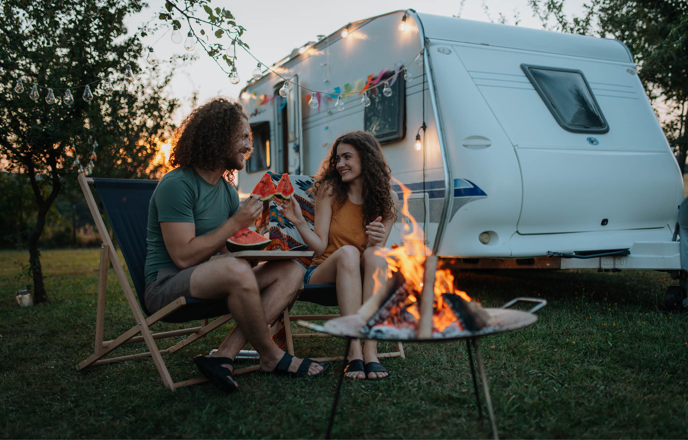 An image of two people sitting down outside a caravan sharing slices of watermelon