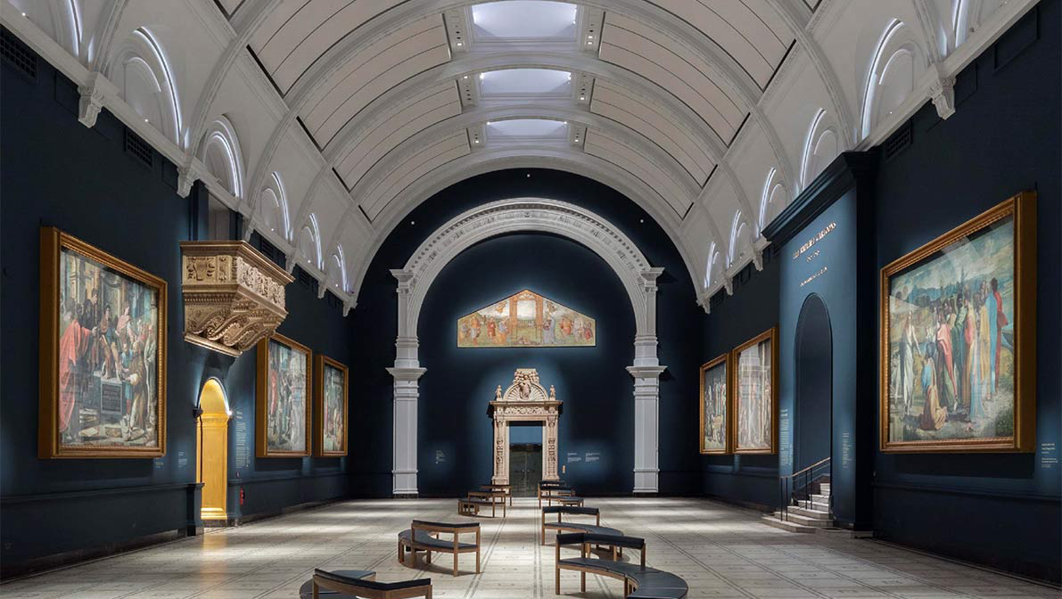 Inside the V and A Museum Raphael Court Avison Young Arts and Culture Specialist Services with blue wall and ornate ceiling arch decoration and historic paintings displayed on the wall next to benches were visitors can view the artwork