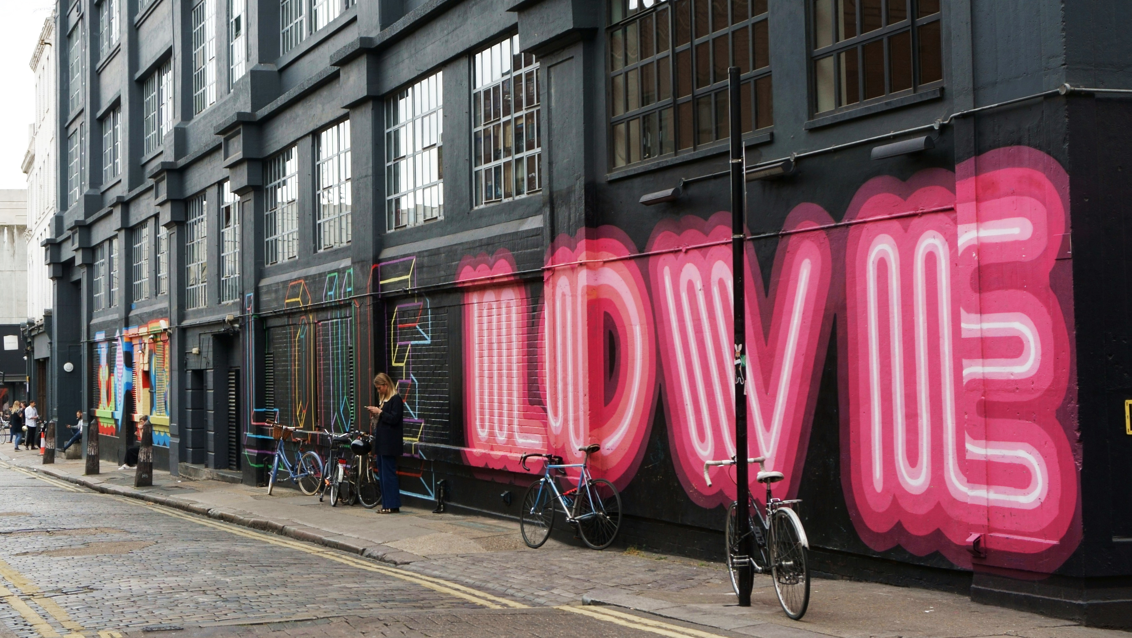 People walking through an urban neighborhood in Shoreditch in London in front of a black painted brick wall with bicycles stood in front of it