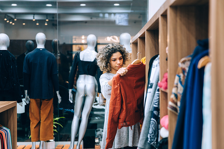 Woman in retail store holding a jacket beside a rail of clothes, behind mannequins in a retail shop window
